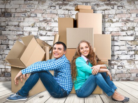 A young man and woman are seated on a light wooden floor in an indoor setting, leaning against a stack of cardboard moving boxes of various sizes, some open with flaps slightly lifted, and others sealed, arranged against a exposed brick wall that features a mix of white and reddish-brown bricks. The man, dressed in a blue checked shirt and jeans, has short dark hair and is smiling at the camera, while the woman, wearing a teal sweater and jeans, has long curly hair and is also smiling, with her arms crossed over her knees. The scene suggests a house relocation or moving process, likely taking place in a residential property in Colliers Wood, near South-West London, with a focus on domestic household items like boxes involved in house removals, supported subtly by the company's service. Natural lighting illuminates the scene, emphasizing the textures of the cardboard boxes, the brick wall, and the casual, friendly atmosphere of the move preparation.
