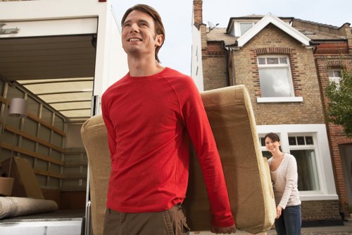 A young man with brown hair, wearing a red long-sleeved shirt and khaki trousers, is outdoors in front of a residential property, carrying a large, beige upholstered armchair with wooden legs. Behind him, a woman with dark hair tied back, dressed in a light striped sweater, is walking towards the house, which features brickwork and white-framed windows, indicating a typical UK suburban street. The scene appears to be part of a house removal or relocation process, with the man lifting the armchair from a vehicle or storage area near the driveway. The environment is bright, suggesting daytime, with natural light illuminating the scene and highlighting the textures of the furniture, brick walls, and clothing. The setting aligns with services offered by Colliers Wood Man with Van, specializing in residential removals within the UK postcode area, likely in or near Colliers Wood, London.