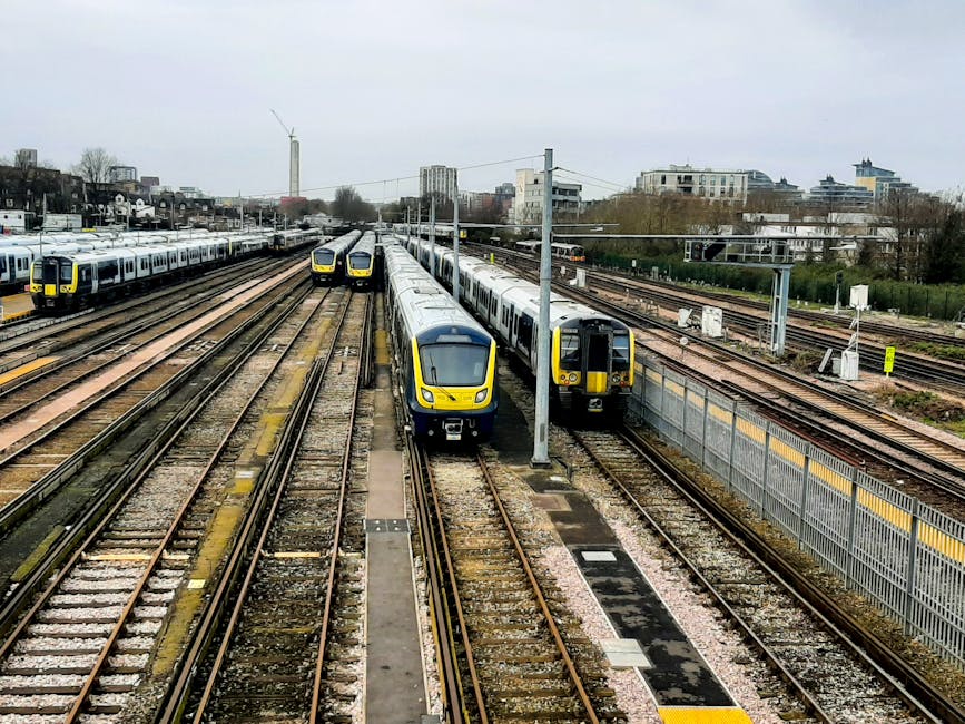 A London Underground train with a red, blue, and silver exterior is stationary at Colliers Wood station platform, which has tactile paving along the edge for safety. The platform features a large wall